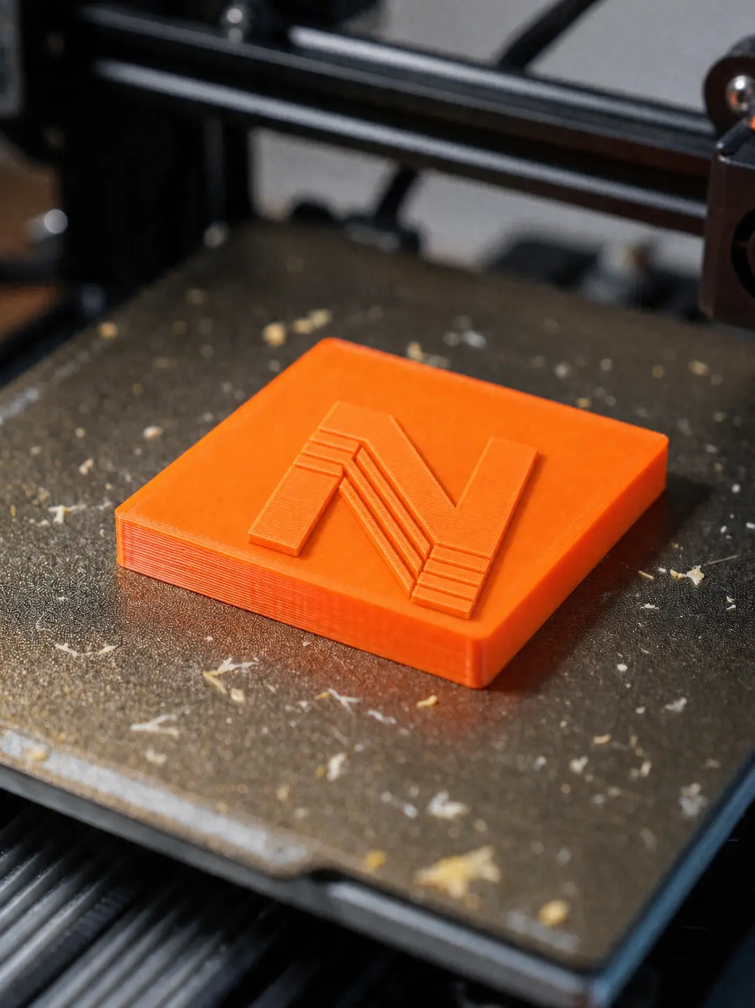 Peça quadrada em PLA laranja com o N da Nave3D em relevo, ainda na mesa da impressora